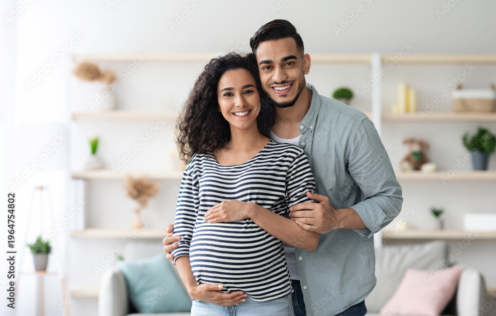 Fototapeta premium Portrait of happy arabic expecting family posing together at home, handsome middle eastern millennial man and beautiful long-haired pregnant lady embracing and smiling at camera, copy space