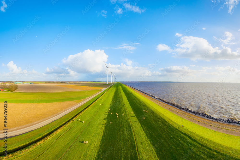 Obraz premium Aerial view of wind turbines on green fields near sea coast and dam. Scenic landscape with wind generators, meadow, ocean and cloudy sky at sunset. Renewable energy and environment from above.