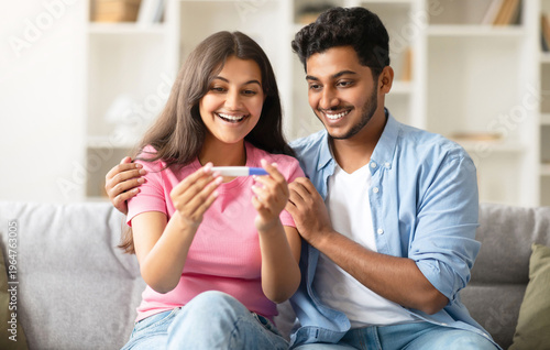 Принти на полотні Excited indian couple looking at positive pregnancy test, smiling and enjoying great news, sitting on couch at home