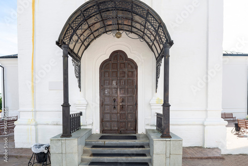 A stone monastery wall with a wooden door facing the street. The door is closed, and the stage is illuminated by sunlight. The door is massive and heavy.