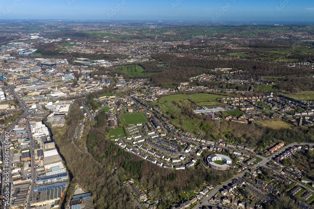 Fototapeta premium Aerial view of the town of Huddersfield in the UK showing terraced housing sports pitch and industrial areas with main road connections and countryside beyond in bright spring daylight conditions