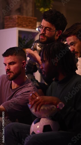 Diverse group of male friends reacting nervously while watching a tense football match on tv at night