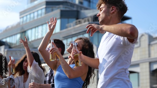 Canvas Print Many young casual people dancing outdoor with drinks at rooftop party on sunny day holding bottles in hands