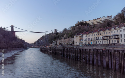 A view of the Clifton Suspension Bridge in Bristol, England, spanning the Avon Gorge, with rows of houses on the hillside and a river in the foreground.