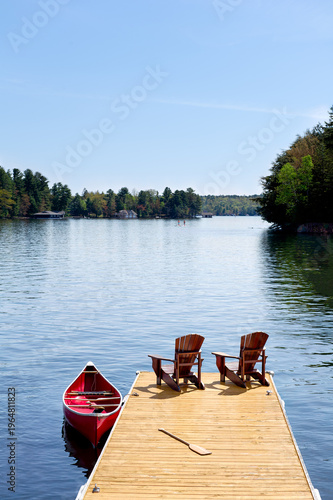 Two Adirondack chairs and a red canoe on a sunny wooden dock in cottage country Ontario, Canada. Paddleboarders enjoy a calm summer lake, vacations, retirement, beauty in nature, tranquility.