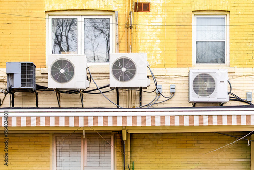 Three mini-split air conditioner condenser units installed side by side on exterior ledge of yellow brick multi-unit building.
