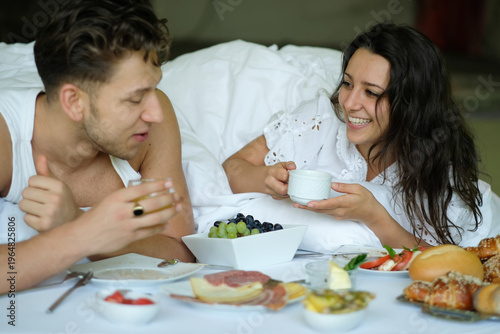 Couple enjoying breakfast in bed smiling and talking while holding cups fresh food on tray relaxed morning scene with cozy atmosphere and intimate connection