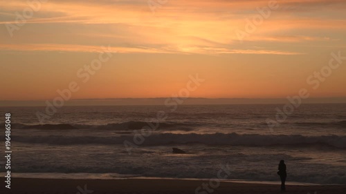 Woman in silhouette walking along shoreline of beach at sunset. Unidentifiable woman on shore.