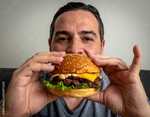 Man holding a cheeseburger with lettuce and sauce, brown sesame bun, casual indoor setting, delicious fast food, appetizing meal.