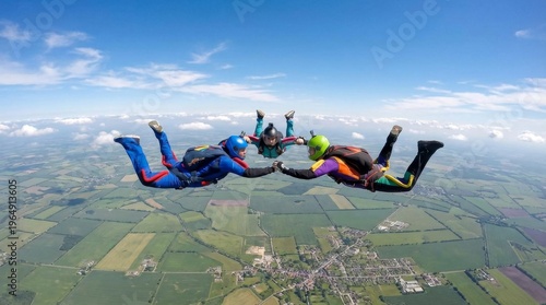Three skydivers in colorful jumpsuits holding hands in freefall above green fields and a small town under a blue sky.