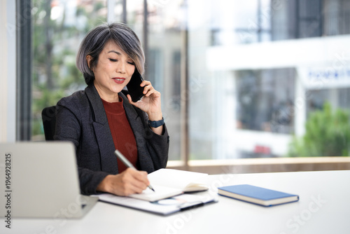 Mature happy joyful confident asian business lady, broker or manager, sitting at table in a modern office, working with laptop, talking to client or employees on smartphone, smiling