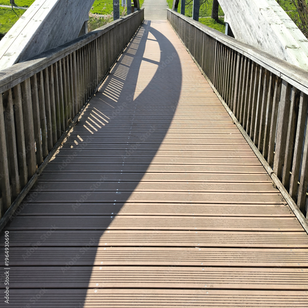 Fototapeta premium Sunlit Wooden Boardwalk Bridge with Geometric Shadow Patterns in a Park Setting