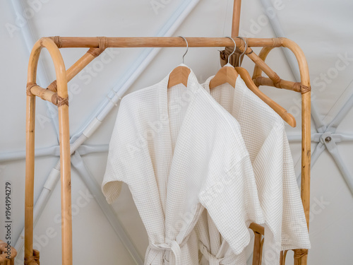 Couple white waffle bathrobes hanging on wooden rack in modern white room, representing spa luxury, hotel and resort amenities, relaxation, wellness lifestyle and minimalist interior design concept.