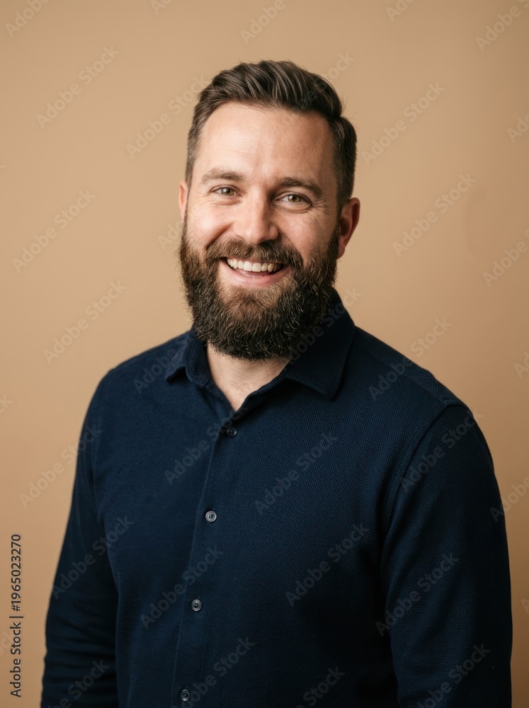 Fototapeta premium A man with a beard and short hair smiles warmly at the camera. He is wearing a dark blue button-down shirt.