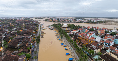 Aerial view of Thu Bon River in Hoi An, Vietnam, with boats lining the riverbanks and people walking along the shore, showcasing the city's vibrant river life.