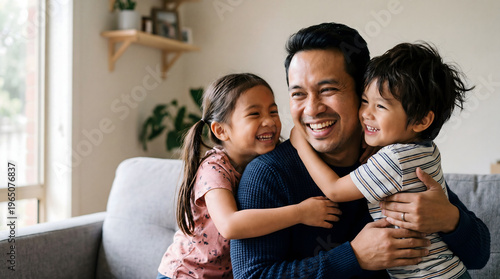 Happy Southeast Asian father laughing while being hugged by his two young children on a sofa at home, candid family bonding moment.