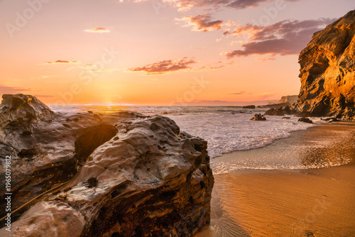 Golden sunset light illuminates rocky shoreline with ocean waves and misty sea spray, creating a peaceful coastal landscape with warm tones, rugged cliffs and natural seaside atmosphere