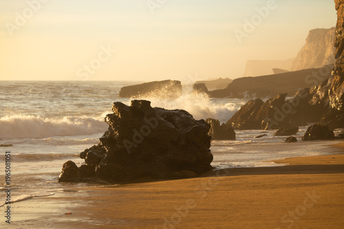 Golden sunset light illuminates rocky shoreline with ocean waves and misty sea spray, creating a peaceful coastal landscape with warm tones, rugged cliffs and natural seaside atmosphere