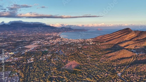 Sunrise aerial over golden ridge bay warm light bathes slopes, distant city and calm sea visible, long shadows