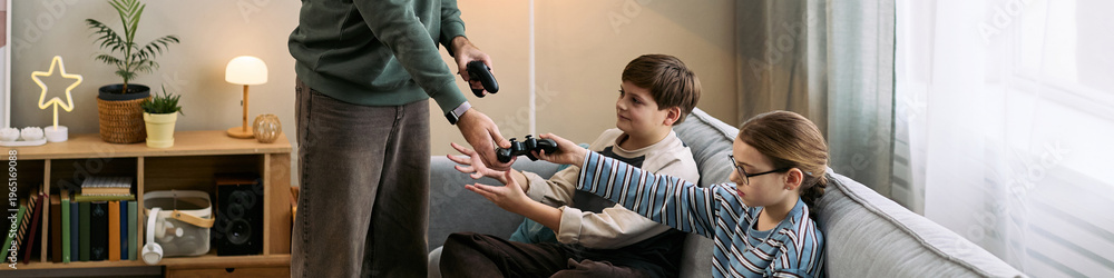 Fototapeta premium Caucasian boy and Caucasian girl sitting on sofa reaching for game controllers from adult man standing nearby, children preparing to play video game together in living room setting