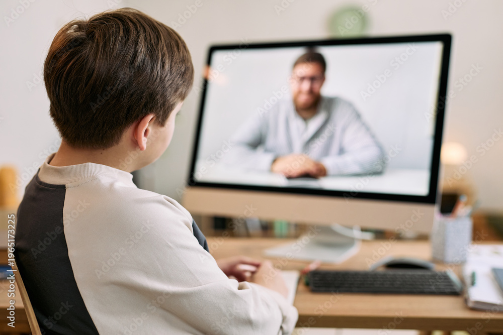 Fototapeta premium Caucasian boy sitting at desk watching middle aged Caucasian man on computer monitor during video call, boy facing screen with hands resting on table, home interior background visible