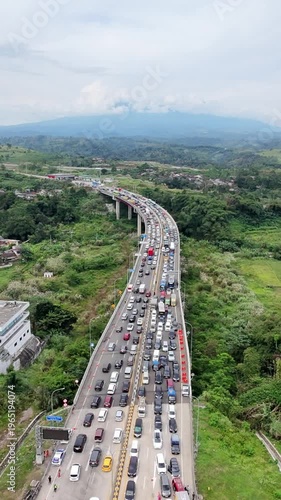 Aerial view of heavy traffic on a curved elevated highway cutting through lush green terrain, with scattered buildings and misty mountains in the distance
