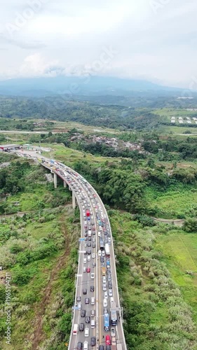 An aerial view of a busy elevated highway curving through lush green countryside, with dense traffic stretching across a scenic landscape of hills and distant mountains