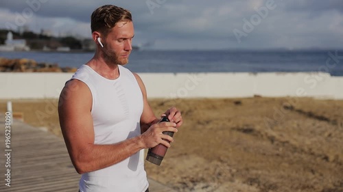 Young strong sporty fit sportsman man wear sports clothes listen ton music in earphones drink protein shake warm up train at sunrise sun over sea beach outdoor on pier seaside in summer day morning