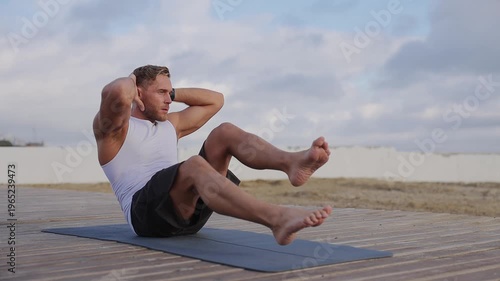 Young strong sporty fit sportsman man wear sports clothes do abdominal abs exercises at floor pump press warm up train at sunrise sun dawn over sea beach outdoor on pier seaside in summer day morning