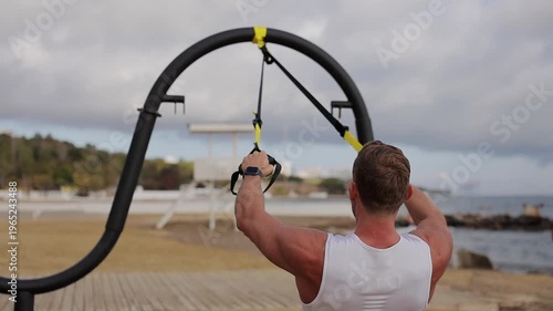 Back rear view young strong sporty athletic fit sportsman man wears sports clothes use trx loops pull up warm up train at sunrise sun dawn over sea beach outdoor on pier seaside in summer day morning