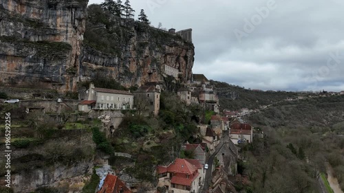 Aerial view of Rocamadour dramatic village and sanctuary buildings clinging to steep rocky cliff face above deep gorge under cloudy sky