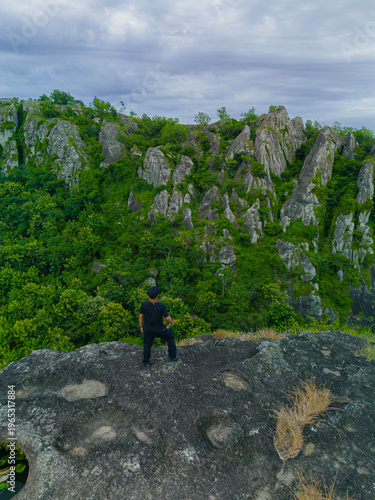 Aerial photo of a male adventurer on a rocky mountaintop. Drone photo of a solo traveler on a pristine & beautiful ancient rocky hilltop with a backdrop of other rocky mountains covered in green trees