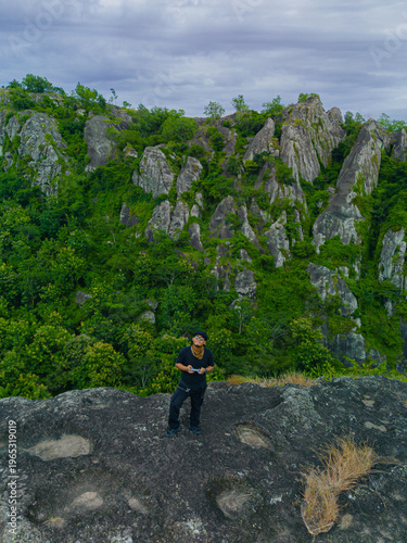 Aerial photo of a male adventurer on a rocky mountaintop. Drone photo of a solo traveler on a pristine & beautiful ancient rocky hilltop with a backdrop of other rocky mountains covered in green trees