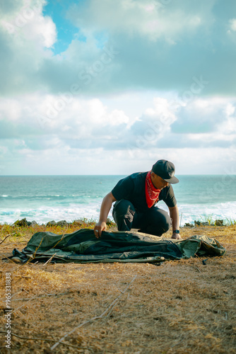 A male adventurer prepares to set up a tent alone by the sea. Solo camping activity on a hilltop near the beach with a view of the vast ocean and clouds. Male adventurer, tent, and ocean all at once.