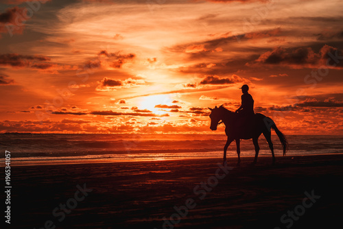 A lone rider on a horse, silhouetted against a breathtaking orange and red sunset sky, walks along the tranquil coastline with ocean waves gently lapping nearby.