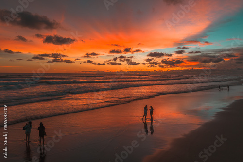 A vibrant sunset casts fiery orange and teal hues across a serene beach where people walk along the wet sand, reflecting the beautiful sky and ocean waves.