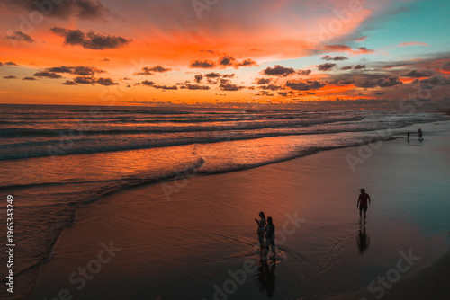 A breathtaking sunset paints the sky in vibrant oranges and reds over a tranquil beach where people enjoy the calming waves and reflections.