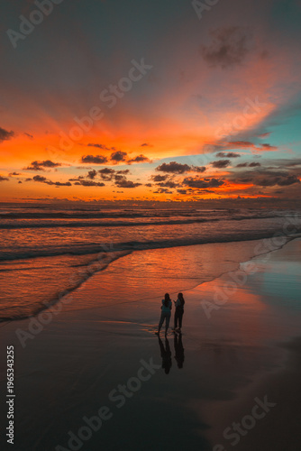 Two figures stand on a reflective beach at sunset, bathed in the dramatic hues of an orange and blue sky over the ocean, creating a picturesque and serene coastal scene.