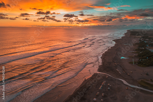 An breathtaking aerial view captures the vibrant orange and teal sunset over a vast ocean with rolling waves, a dark sandy beach dotted with people and umbrellas, and a tranquil coastline.