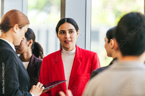 Professionals converse at a networking event; a woman in a red blazer speaks with colleagues near large windows while another uses a tablet.