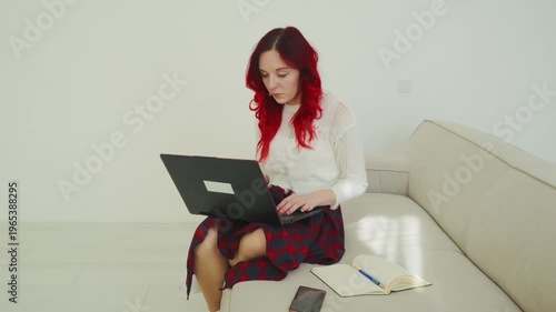Young Woman with Vibrant Red Hair Working on Laptop Sitting on a Sofa in a Minimalist White Interior