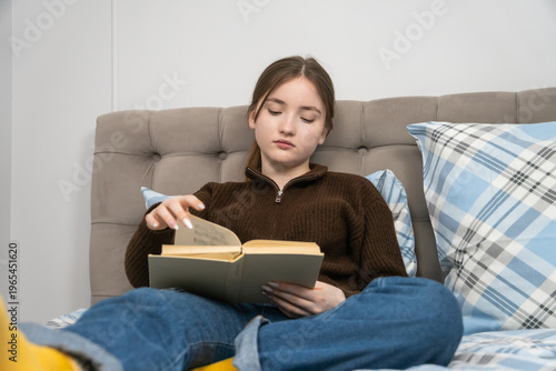 Teen girl relaxing on a bed in natural light, comfortably turning pages of a book while studying at home - cozy, focused moment of quiet learning and personal growth