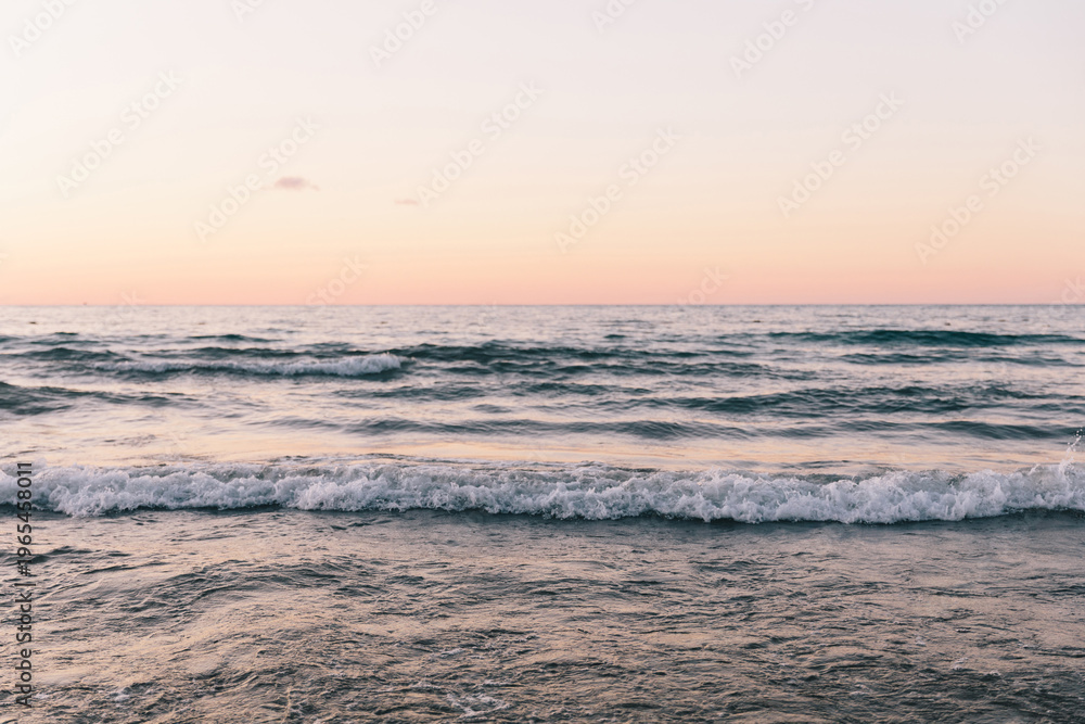 Fototapeta premium Calm ocean waves with white foam roll towards the shore under a soft pink and peach twilight sky. The minimalist seascape captures a tranquil and meditative atmosphere during the golden hour.