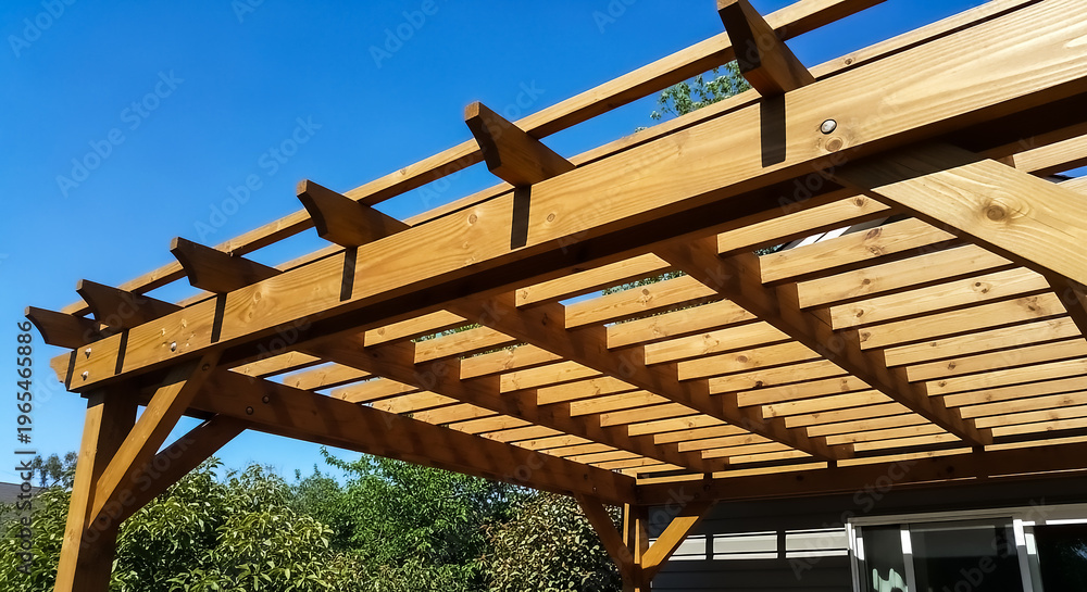 Fototapeta premium A well-constructed wooden pergola with visible beams and rafters, set against a bright blue sky and lush greenery backdrop.