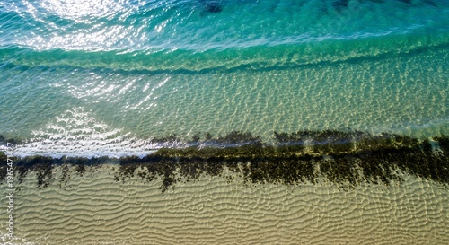 Breathtaking aerial view of clear turquoise ocean waves gently washing over a sun-drenched sandy beach, revealing intricate seabed patterns and a tranquil coastal landscape