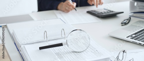 Magnifying glass and calculator are on the table over financial documents opposite female accountant analyzing data, reviewing reports, binders and laptop on a white desk. Audit and taxes concept
