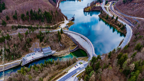 Aerial view of the Staudamm Klaus dam reflecting the sky's hue, nestled amongst autumnal trees and curving roads, Klaus an der Pyhrnbahn, Oberosterreich, Austria.
