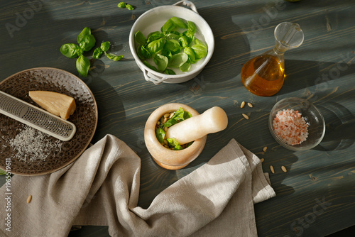 Traditional Genovese pesto preparation with fresh ingredients on wooden background. High angle shot of marble mortar with pestle, fresh basil, pine nuts, olive oil and parmesan cheese with grater for