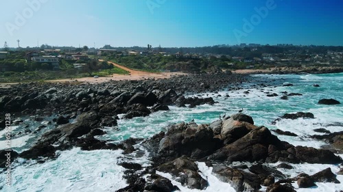 Slow forward flight over dark jagged rocks with seabirds perched and turquoise waves crashing revealing sandy cove and coastal residential town beyond. El Tabo, Chile