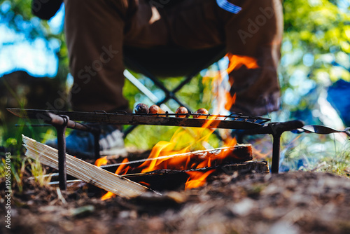 Close-up of food grilling over a bright orange campfire flame with a person sitting in a folding chair behind.
